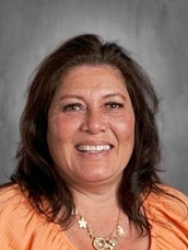 A woman with shoulder-length brown hair smiling, wearing an orange top and a necklace, against a gray background.