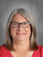 A woman with shoulder-length gray hair and glasses smiles in front of a gray background, wearing a red shirt.