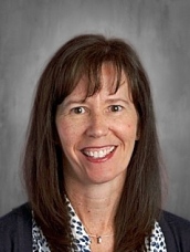 Smiling woman with shoulder-length brown hair, wearing a patterned blouse and dark jacket.