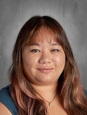 A woman with long, layered hair and a slight smile is posed against a gray background. She wears a blue top and a necklace.
