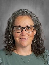A person with curly hair and glasses, wearing a green top, smiles at the camera against a neutral background.