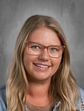 A person with long blonde hair and glasses smiles in front of a gray background.