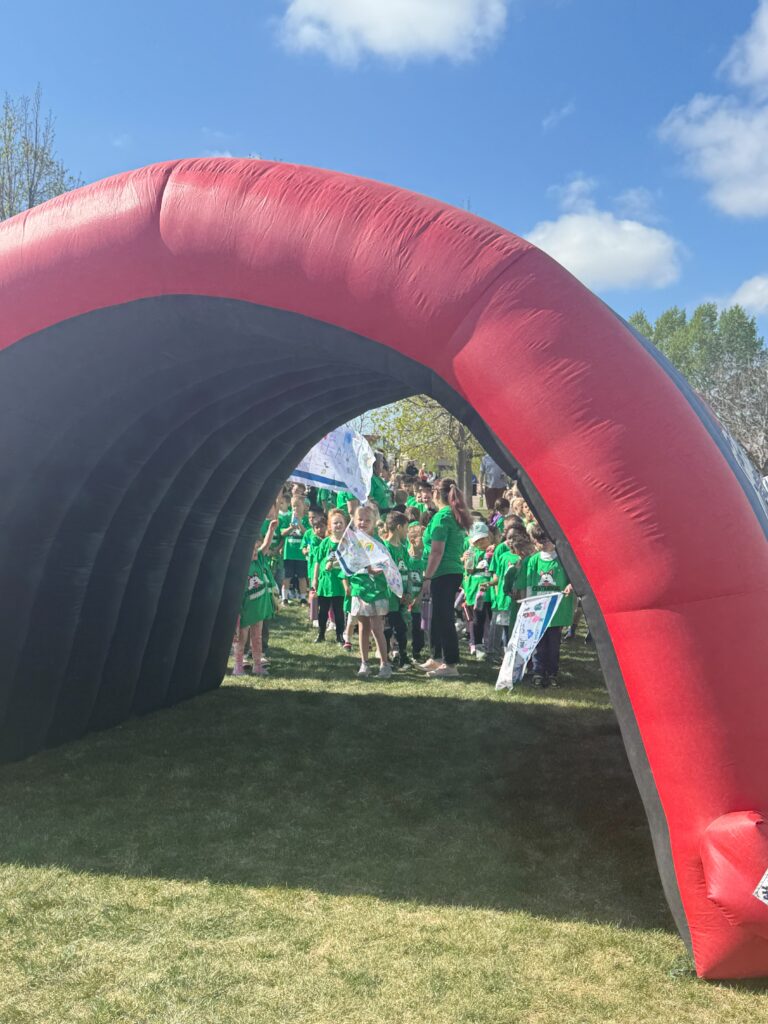 A group of children in green shirts gathers under a large inflatable arch on a sunny day.