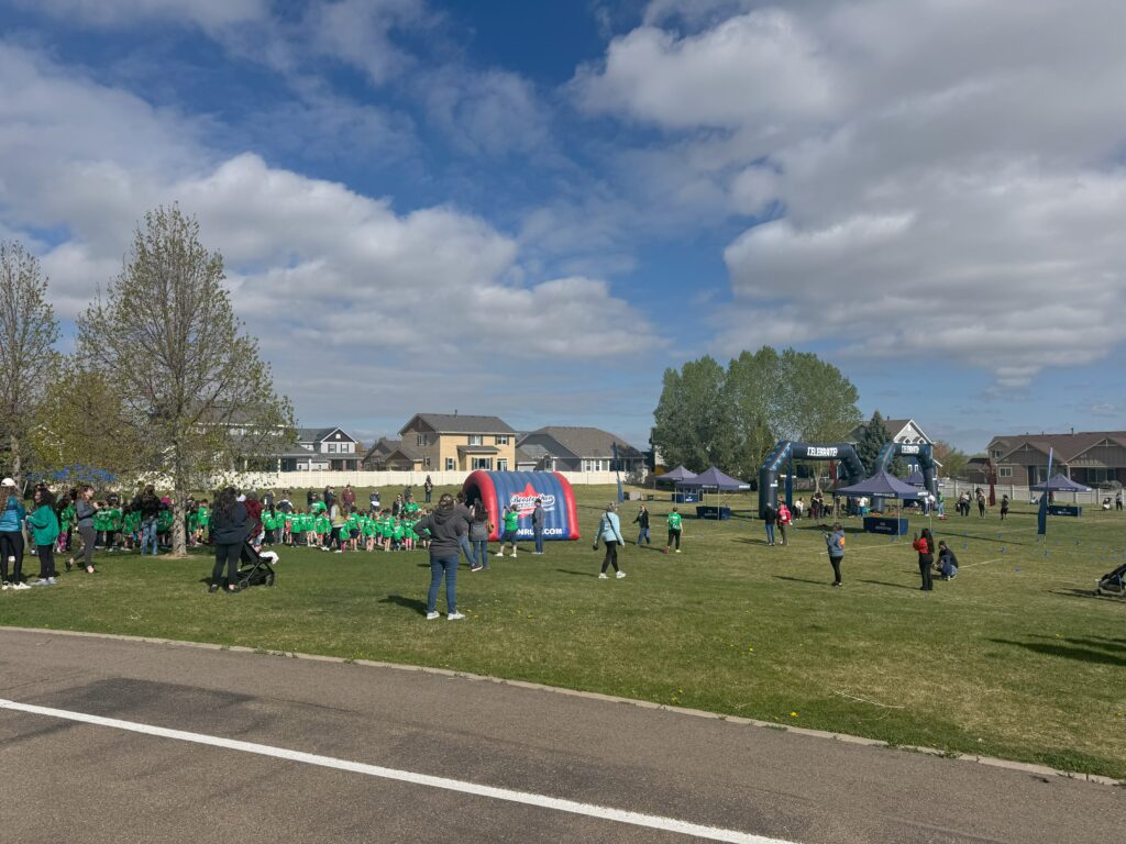 A community event in a park with groups of children in green shirts, tents, and an inflatable archway under a blue sky.