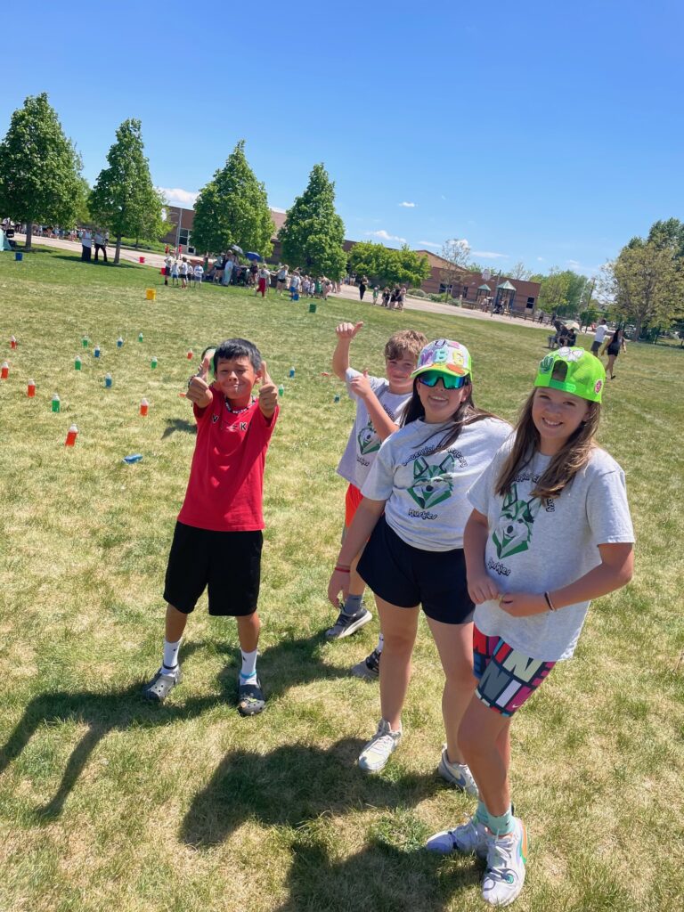 Four children pose cheerfully outdoors on a sunny day, surrounded by colorful cones in a grassy field.