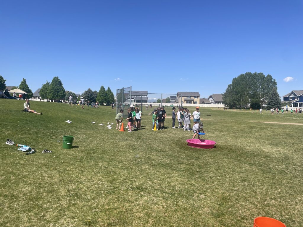 A sunny outdoor field filled with children participating in activities, with houses and trees in the background.
