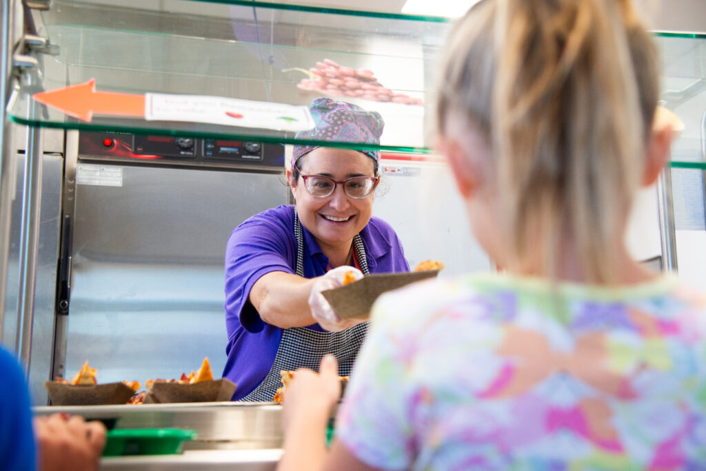 A smiling worker hands food to a child at a serving counter in a school cafeteria. Colorful decor is visible.