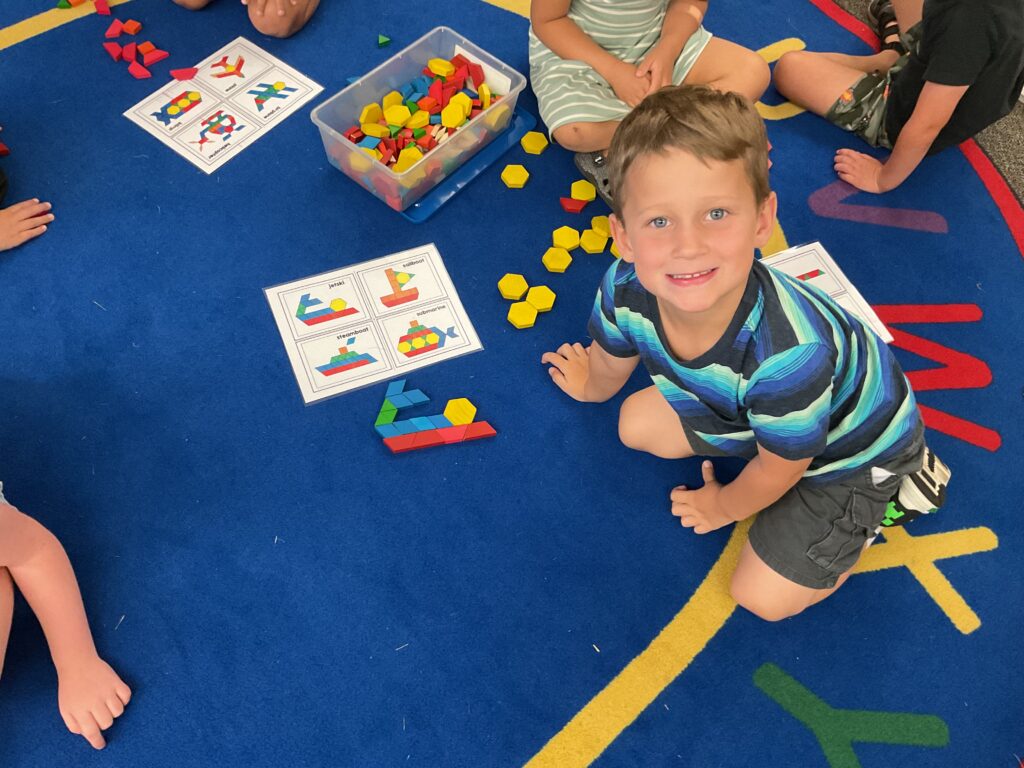 A young boy smiles while playing with colorful building blocks on a blue carpet, surrounded by peers and activity sheets.