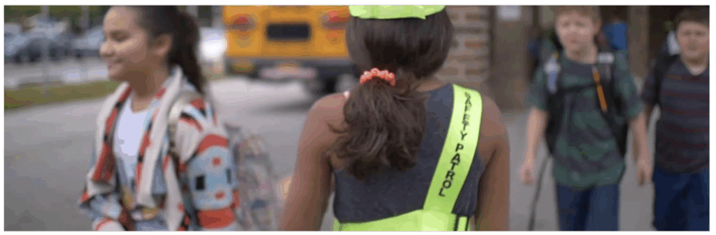 A child wearing a "Safety Patrol" vest walks alongside peers near a school bus and entrance.