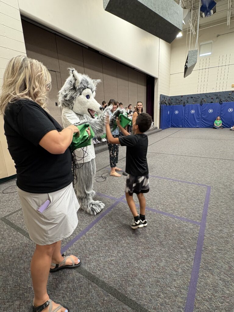 A child interacts with a wolf mascot while adults and other kids watch in a gymnasium setting.