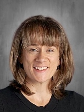 A woman with brown hair and bangs smiles, wearing a black shirt and large earrings, against a neutral background.