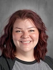 Smiling woman with wavy, shoulder-length reddish-brown hair, wearing a gray shirt with black sleeves.