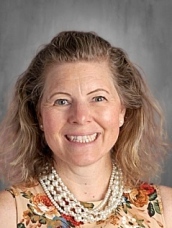 Smiling woman with wavy hair, wearing a floral dress and pearl necklace, set against a gray background.