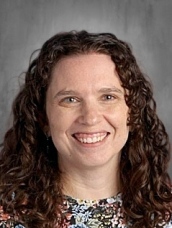 Smiling woman with curly brown hair wearing a floral shirt, set against a neutral background.