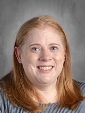 Smiling woman with long red hair, wearing a striped shirt, against a gray background.