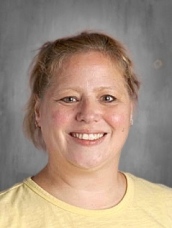 Smiling woman with short, light brown hair wearing a yellow shirt, set against a gray background.