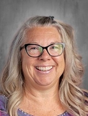Smiling woman with long, wavy gray hair and glasses, wearing a colorful top, against a neutral background.