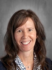 Smiling woman with long, wavy brown hair, wearing a patterned blouse, set against a neutral background.