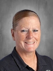 A smiling person with short brown hair wearing a dark shirt, set against a gray background.