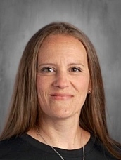 A woman with long brown hair smiles, wearing a black shirt, against a neutral background.