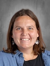 Smiling woman with medium-length brown hair, wearing a denim shirt and earrings, set against a gray background.