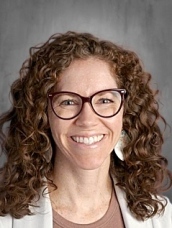 Smiling woman with curly hair and glasses, wearing a light blazer, against a neutral background.