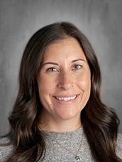 Headshot of a smiling woman with long, wavy brown hair, wearing a gray sweater, against a neutral background.