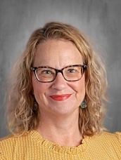 A woman with curly hair, glasses, and red lipstick smiles in front of a gray background, wearing a yellow blouse.