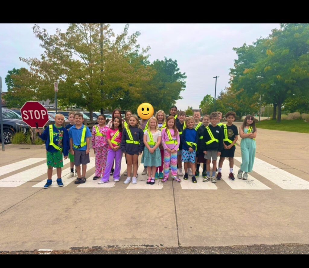 A group of children in bright vests stands on a crosswalk, smiling, with a stop sign nearby and trees in the background.