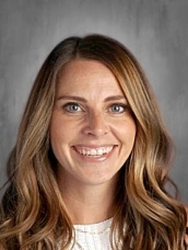 A smiling woman with long, wavy brown hair and blue eyes, wearing a white top, against a gray background.