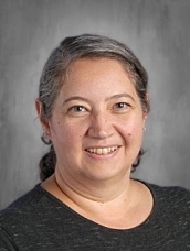 A woman with gray hair tied back, smiling, wearing a dark shirt, set against a neutral background.