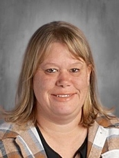 A smiling woman with long, light brown hair, wearing a plaid shirt, against a gray background.