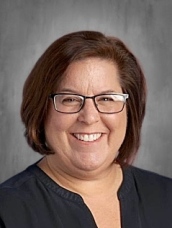 Smiling woman with short brown hair and glasses, wearing a dark top, against a gray background.