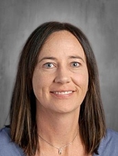 Headshot of a woman with long brown hair, smiling, wearing a blue top against a gray background.