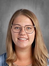 Smiling young woman with long blonde hair and glasses, wearing a denim jacket, against a gray background.