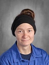 A person with a bun hairstyle wearing a black headband and a blue shirt, smiling against a gray background.