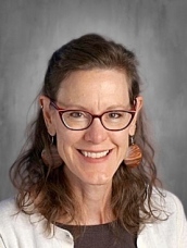 A smiling woman with glasses and earrings, wearing a white cardigan over a dark top, against a gray background.