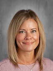 A woman with shoulder-length blonde hair smiles at the camera, wearing a light pink sweater. Background is gray.