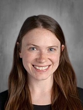 A smiling woman with long brown hair and blue eyes, wearing a black shirt, against a gray background.