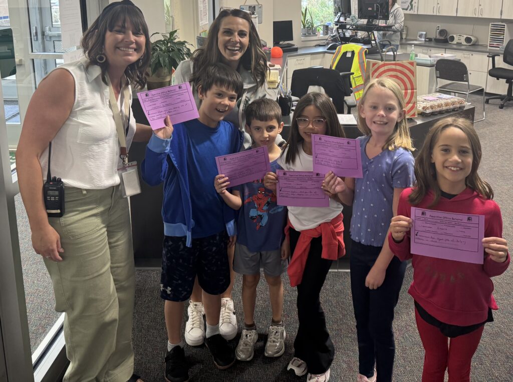 Two teachers and five children smile while holding pink certificates in a school office setting.