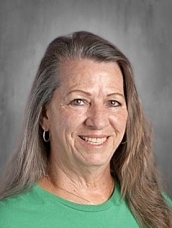 Smiling woman with long gray hair wearing a green shirt, set against a gray background.