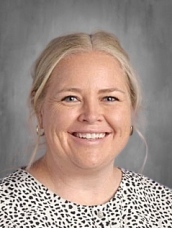 Smiling woman with blonde hair, wearing a spotted blouse, against a gray background.