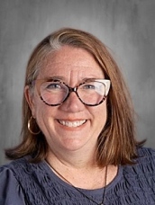 A woman with shoulder-length brown hair and glasses smiles at the camera, wearing a blue top against a gray background.