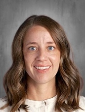 A smiling woman with long, wavy brown hair and blue eyes, wearing a light-colored top, against a gray background.