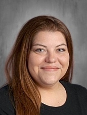 A smiling woman with long brown hair, wearing a black shirt, poses for a professional headshot.