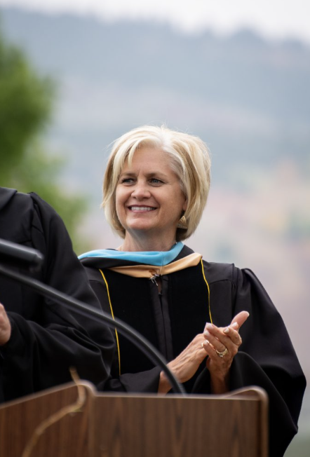 A smiling woman in academic regalia stands at a podium, applauding during a graduation ceremony outdoors.