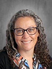 Smiling woman with curly gray hair and glasses, wearing a colorful patterned shirt, against a gray background.