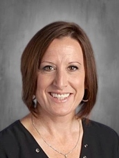 Headshot of a woman with shoulder-length brown hair, wearing a black top and silver jewelry, smiling against a gray background.