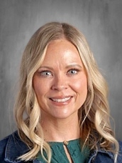 Smiling woman with long, blonde hair, wearing a green top and denim jacket, against a gray background.