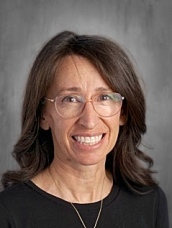Smiling woman with long brown hair and glasses, wearing a black shirt, against a gray background.